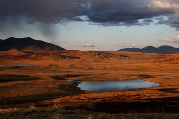Colorful sunset above highland steppe shore of lake with dry yellow grass on the background of rocky mountains under storm sky