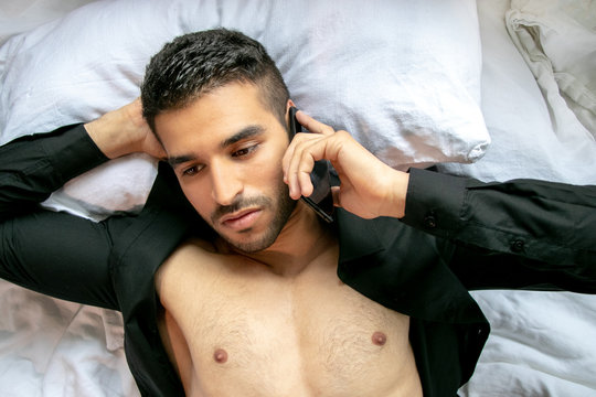 Handsome man with open black shirt lies in bed using mobile cell phone