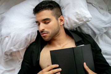 Man in bed with open shirt and pecs sleeps reading hardback book