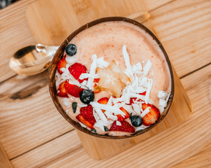 Beautiful pink strawberry smoothie bowl with coconut shavings, banana, blueberries and star fruit on the wooden table with gold spoon.