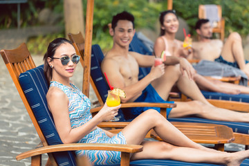 Young and beautiful Asian woman smiling while enjoying a delicious pineapple cocktail with her friends at a trendy swimming pool 