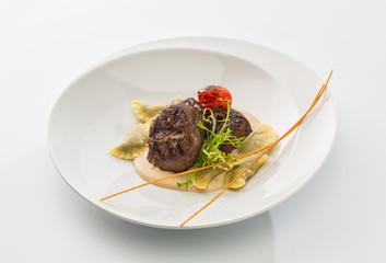 two pieces of roasted meat with sauce and ravioli on a plate isolated on white background