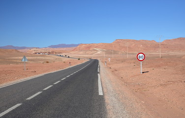 Asfaltowa szosa przez pustynię, w dali jedzie nią sznur samochodów, także dużych camperówk, znak na poboczu z ograniczeniem prędkości, słupy elektryfikacyjne, na horyzoncie wzgórza i rysujące się góry