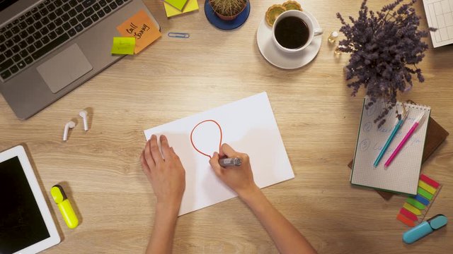 Girl draws image of a "idea bulbs" on a sheet of paper, sitting at work table, first-person view of hands.