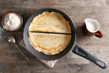 Frying pan with tasty thin pancakes and ingredients on wooden table