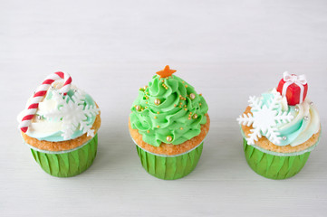 New year or christmas green cupcakes with whipped cream, decorated with a christmas tree, candy cane, a snowflake, a red box, silver and gold confectionery balls on white wood table.