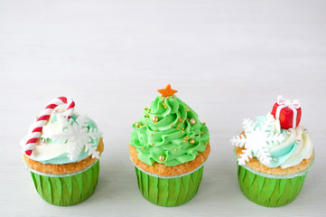 New year or christmas green cupcakes with whipped cream, decorated with a christmas tree, candy cane, a snowflake, a red box, silver and gold confectionery balls on white wood table.