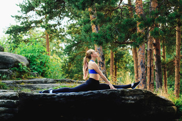 Girl sitting on the splits outdoors on a rock in the forest. Yoga nature concept.