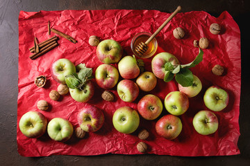 Ripe organic gardening green red apples with leaves, nuts, cinnamon and jar of honey on red bright crumpled paper over dark texture background. Flat lay, space. Autumn harvest.