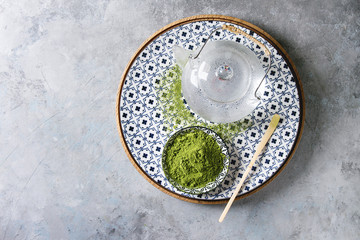 Ingredients for making matcha drink. Green tea matcha powder in ceramic bowl, traditional bamboo spoon on decorative plate, glass teapot over grey texture background. Flat lay, space