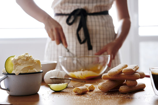 Stack of ladyfingers and cup of mascarpone on wooden table for preparing Tiramisu