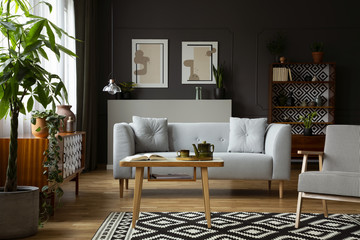 Wooden table and armchair on patterned carpet in retro flat interior with grey sofa and posters. Real photo