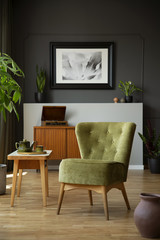 Green chair next to wooden table in dark living room interior with poster and plants. Real photo