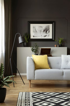Yellow cushion on grey sofa next to lamp in scandi living room interior with poster. Real photo