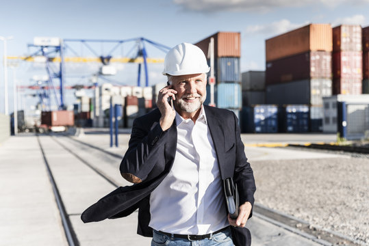 Businessman at cargo harbor, wearing safety helmet, using smartphone