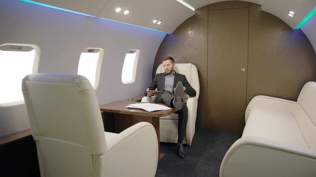 Elegant investor expert analyst businessman is having time with tablet sitting in private plane, successful person using device, looking out window during business trip indoors. Concept: entrepreneur