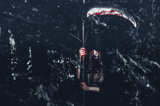 Female demon witch with bloody reaper standing in front of mystery forest background. Halloween and Religious concept. Demon angel and Satan theme.
