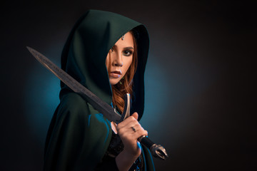 Mysterious young woman keeping sharp knife, wearing green cape with a hood.