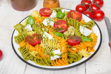 Kuchnia włoska - Sałatka z kolorowego makaronu, pomidorów koktajlowych, sera feta i świeżej bazylii na białym drewnianym tle.