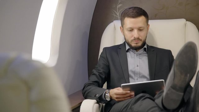 Confident chief banker director businessman sits down in airplane chair, takes tablet, looks out window, attractive bearded guy in suit has good time in luxurious interior during flight. Concept