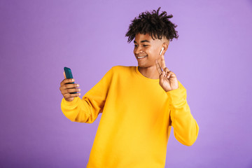 Portrait of young african american man smiling and taking selfie on smartphone, wearing bluetooth earpod, isolated over violet background