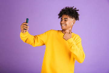 Portrait of teenage african american man smiling and taking selfie on smartphone, wearing bluetooth earpod, isolated over violet background