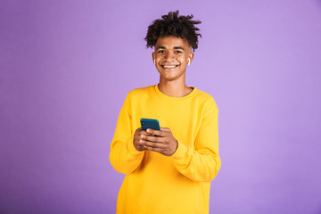 Portrait of teen african american man smiling and holding smartphone, wearing bluetooth earphones,...