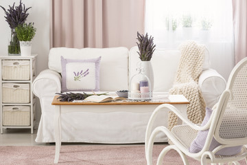 Wooden coffee table with pastel candles, fresh lavender and open book in real photo of bright sitting room interior with white couch