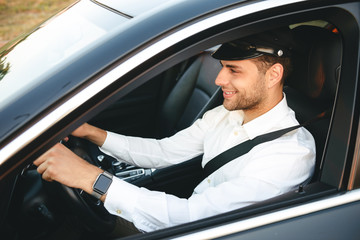 Portrait of happy european man taxi driver wearing uniform and cap, driving car fastening seat belt