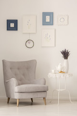 Grey armchair standing next to metal end table with jug and tea cup, french cookies and fresh lavender in white room interior with posters on wall