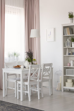 Real photo of dining room interior with chairs and white table with lavender and breakfast, window with drapes and rack with decor