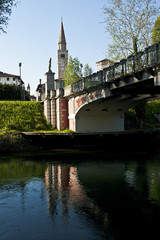 The Adam and Eve bridge over the Noncello river in Pordenone. In the background the bell tower of the Cathedral of San Marco. Italy.