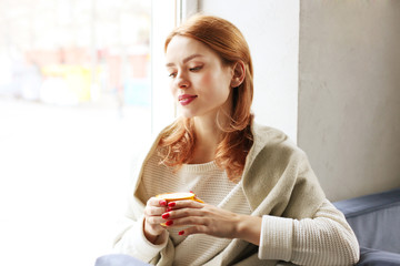 Beautiful redhead woman, sitting near coffee shop window holding yellow cup of hot beverage, wearing winter autumn season white knitted sweater & big loose scarf on shoulders. Background, close up.
