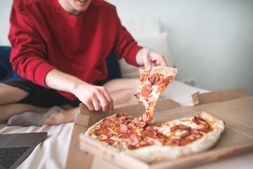 Young man in a red sweatshirt sits at home on a bed and takes an appetizing piece of pizza from the box. Men's hands take a delicious piece of pizza. The teenager is going to eat a piece of pizza - obrazy, fototapety, plakaty