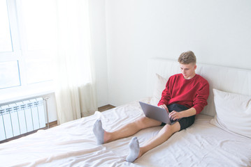 Young man sitting at home in a bright room on a bed with a laptop on his lap and working. Work at home on the Internet. Freelancer works on a laptop in his bed room