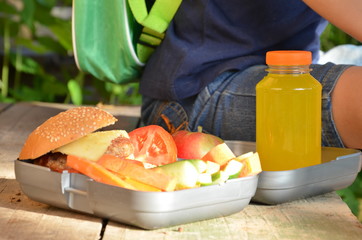 Cute schoolboy eating outdoors the school from plastick lunch boxe. Healthy school breakfast for child. Food for lunch, with sandwiches, fruits, vegetables (carrots cucumber tomato) , and juice