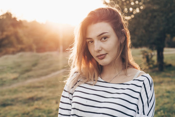 Fototapeta premium Young woman relaxing in summer sunset sky. Portrait of Beautiful Young Woman Backlit at Sunset Outdoors. Soft warm sunny colors.