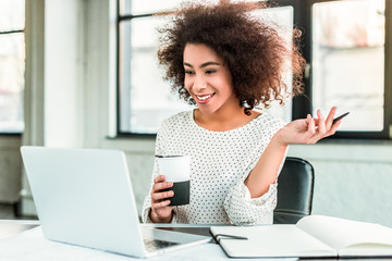 happy african american businesswoman holding cup of coffee and looking at laptop in office
