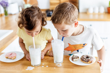 Children eating donuts and drinking milk on the white kitchen at home. Child is having fun with donuts. Tasty food for kids. - Powered by Adobe