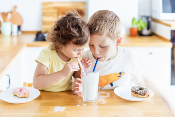 Children eating donuts and drinking milk on the white kitchen at home. Child is having fun with...
