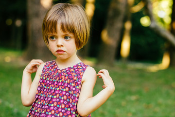 Close up portrait of sad pensive cute baby girl in summer park. Child touch their shoulders outdoor in green garden, blurred background.