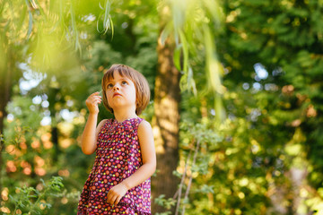 Cute baby playing outdoor in summer park alone. Three-year-old child in colorful dress on green background. Curious baby girl researcher touching a leaf with finger. Question emotion in face