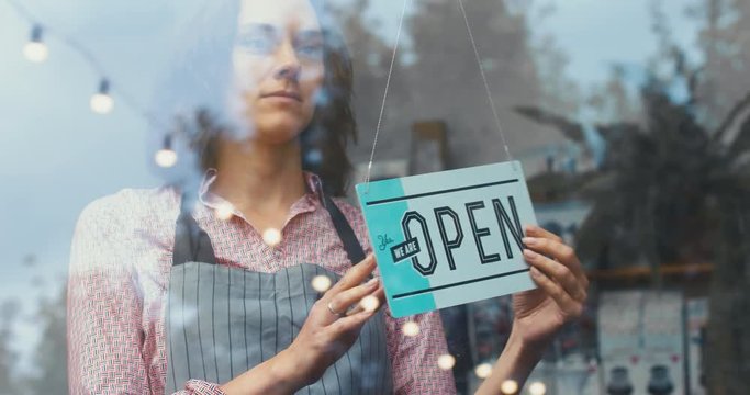 Caucasian female turning sign from open to closed on the entrance door or window of her small cafe and taking off apron. 4K UHD 60 FPS SLOW MOTION