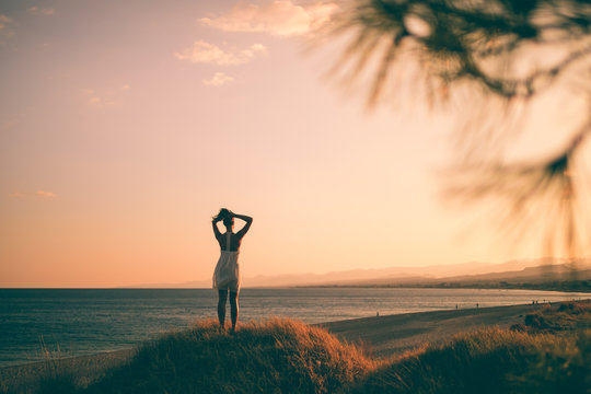 Ragazza solitaria con vestito bianco in riva all'oceano guarda l'infinito concentrata e pensando al futuro durante un tramonto rosso fuoco.