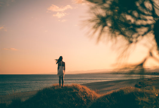 Ragazza solitaria con vestito bianco in riva all'oceano guarda l'infinito concentrata e pensando al futuro durante un tramonto rosso fuoco.