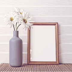 A bouquet of three white chamomiles is in a gray vase in the form of a bottle. Nearby there is an empty wooden frame. White wooden background.
