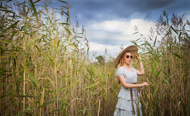 A blonde girl in a white dress with a blue print and a straw hat is walking among the reeds.