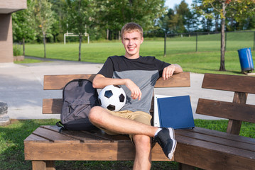 Happy teenager sitting on a bench with a soccer ball and his school supplies.