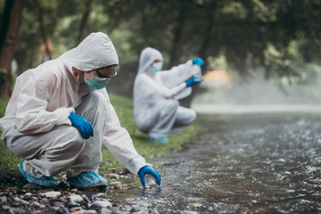 Two scientists in protective suits taking water samples from the river. - obrazy, fototapety, plakaty
