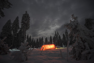Winter camping, night, shining red tent on snow, clouds, forrest. Night shot, long exposure, sleeping on snow in the outdoors.